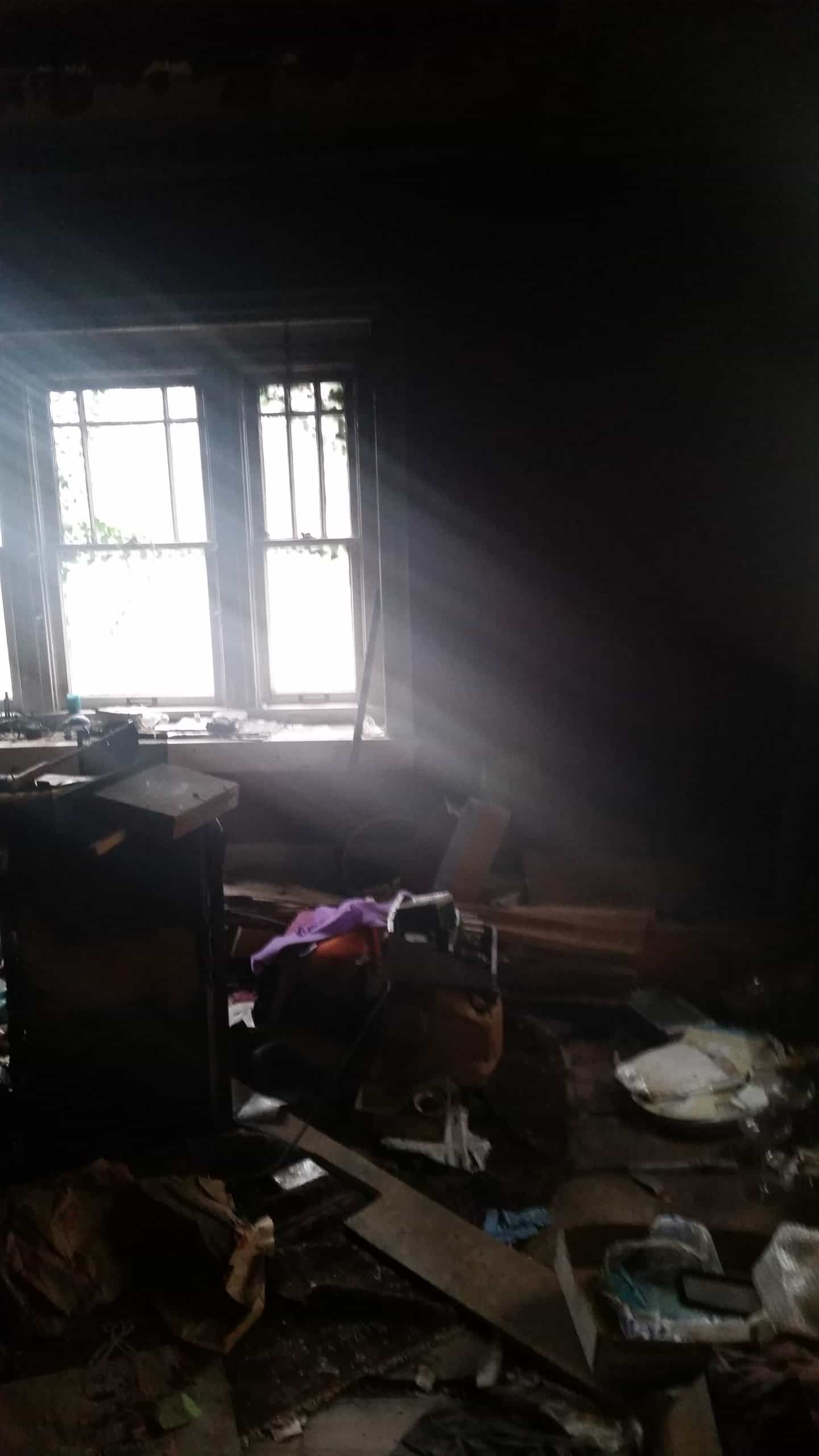 Interior of a dark house with debris piled on the floor and a window letting light in.